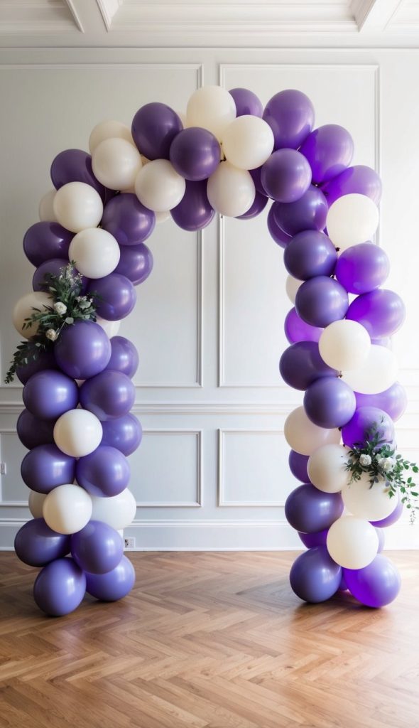 A decorative arch made of purple and white balloons with green foliage and white flowers, set indoors against a white paneled wall and hardwood floor.