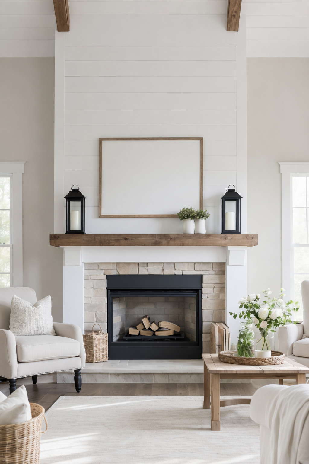 A cozy living room with a white shiplap wall and a stone fireplace. The mantle features decorative black lanterns and small potted plants. Light-colored chairs with pillows flank the fireplace, and a wooden coffee table displays a basket with flowers. A beige rug covers the hardwood floor.
